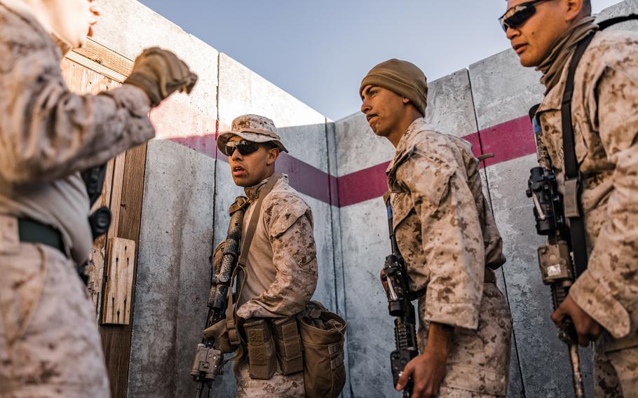 Four Marines armed with guns stand by a wall in front of a wooden door.