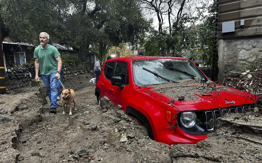 A man walks a dog past a red car submerged in mud up to the wheels.