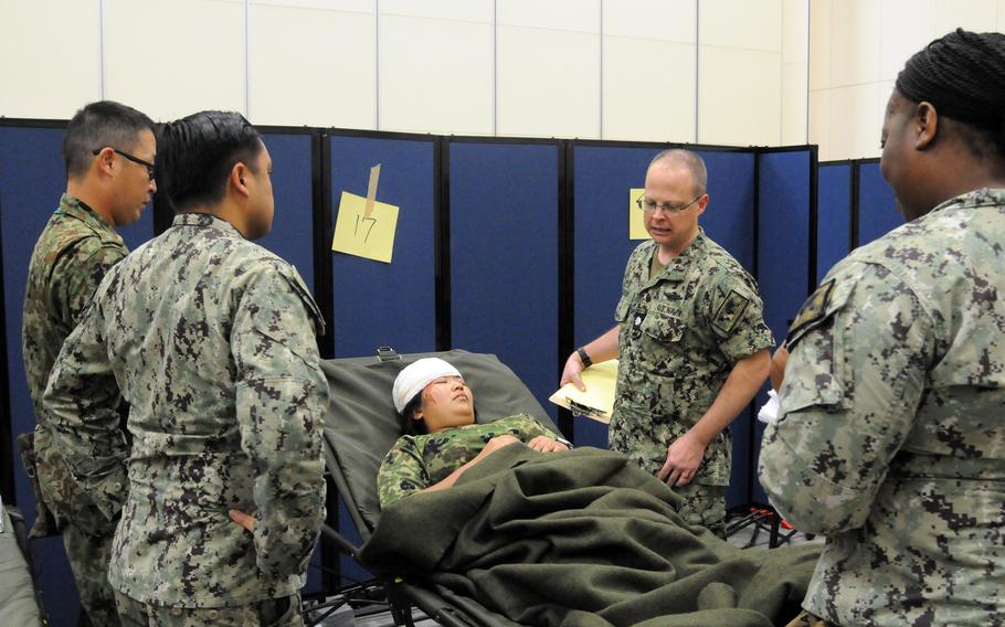 Military service members in uniform stand above a patient with simulated injuries laying on a stretcher.