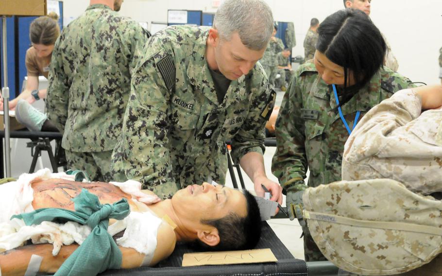 Two military service members dressed in uniform stand over a man with simulated injuries laying on a stretcher.