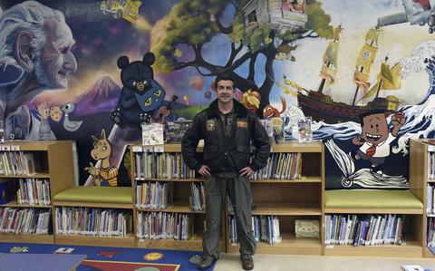 A man in a flight suit and bomber jacket stands in front of a large painted mural on a wall above book shelves.