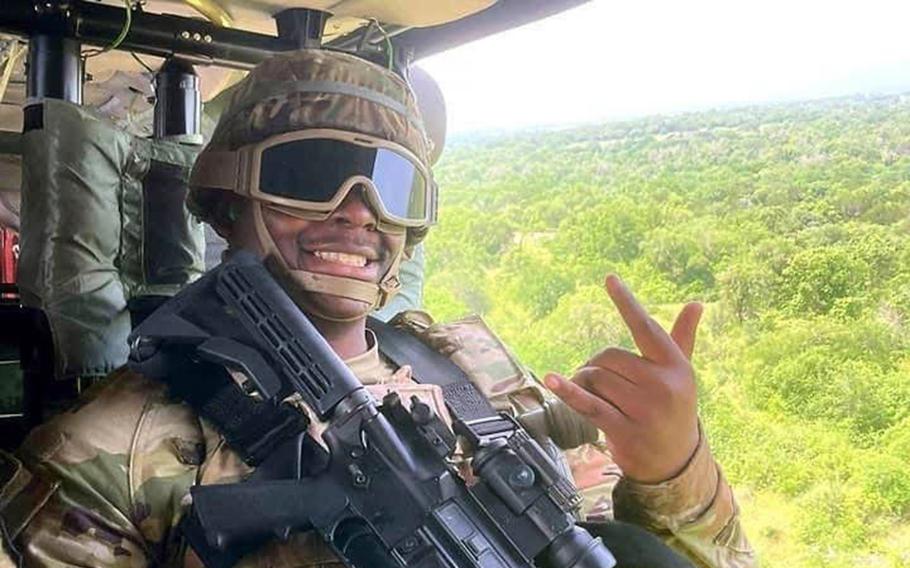 A man dressed in an Army uniform and wearing a gun rides in a helicopter and flashes a backwards I love you hand signal at the camera.