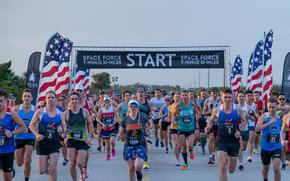 Dozens of runners are seen heading towards the camera, in front of the start line for the Space Force’s annual 10-mile marathon.