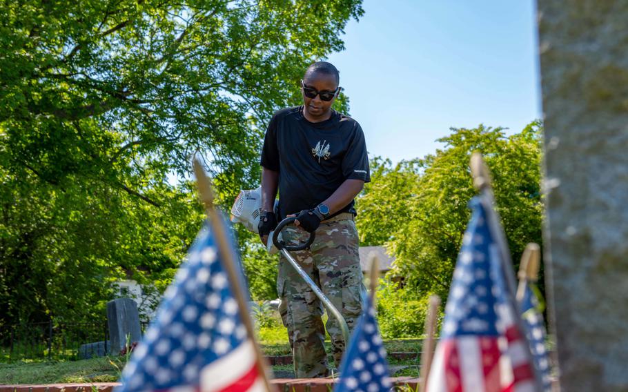 A man in a T-shirt and camouflage pants cuts grass with American flags in the foreground.