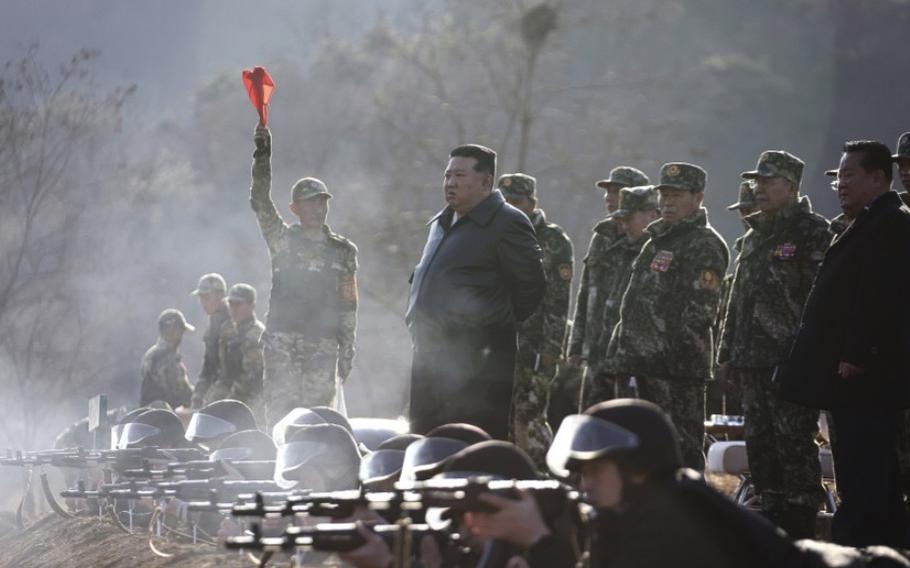 Kim Jong Un stands behind a row of North Korean troops aiming weapons and is flanked by more troops.