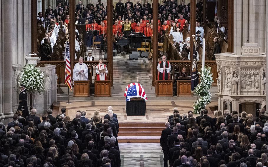 A casket draped with a U.S. flag sits on a pedastal in the middle of a cathedral as mourners stand during a state funeral.