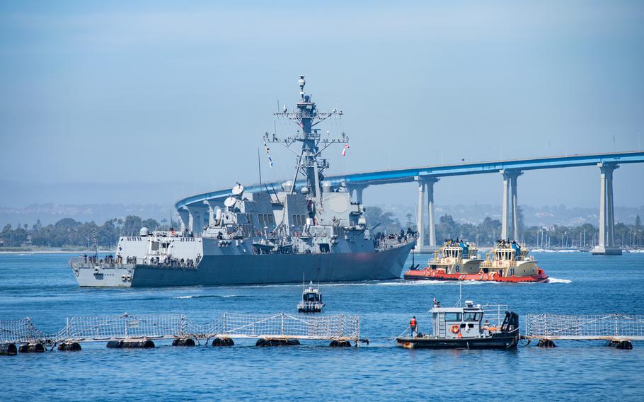 USS Spruance heads out to sea. A bridge in San Diego Bay is in the background, and support boats are nearby.