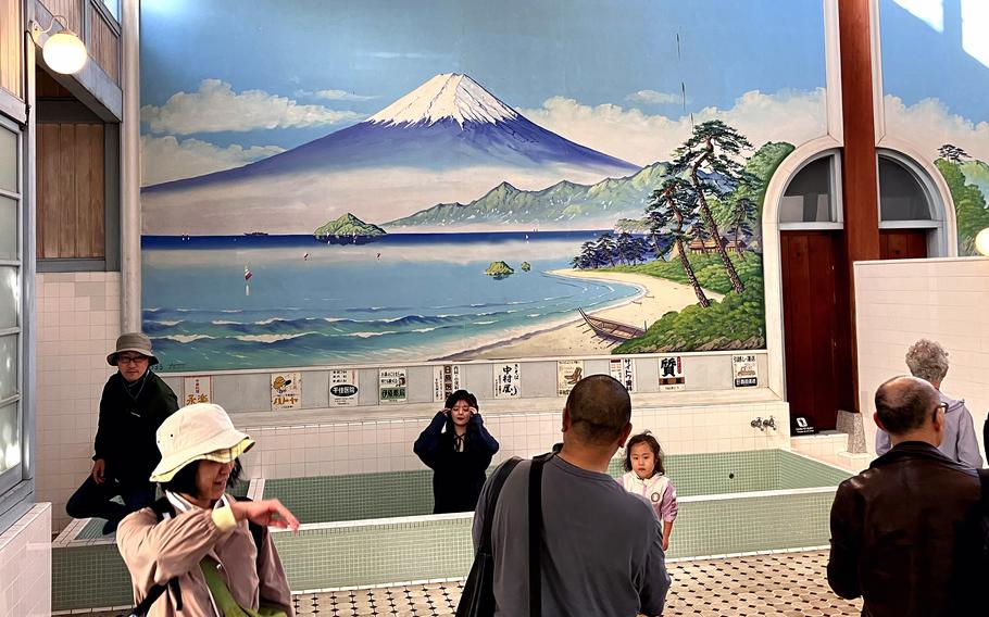 A mural showing a painting of Mount Fuji and a lake adorns a wall with people milling around in front of it.