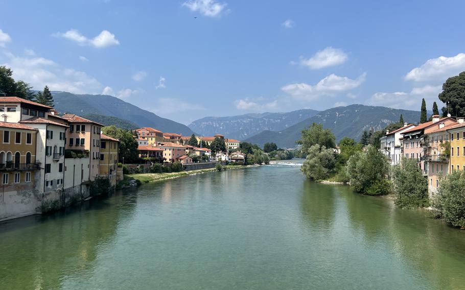 A river lined on each side with buildings in Bassano del Grappa, Italy, stretches on to the horizon with the Alpine mountains in the background on a sunny day.