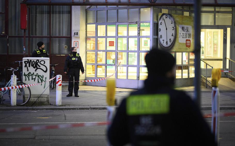 The back of a police officer is seen out-of-focus in the foreground, while an in-focus background shows another police officer and the front of a supermarket.