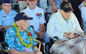 Two elderly men sit side by side in wheelchairs, each wearing a hat that says Pearl Harbor survivor.