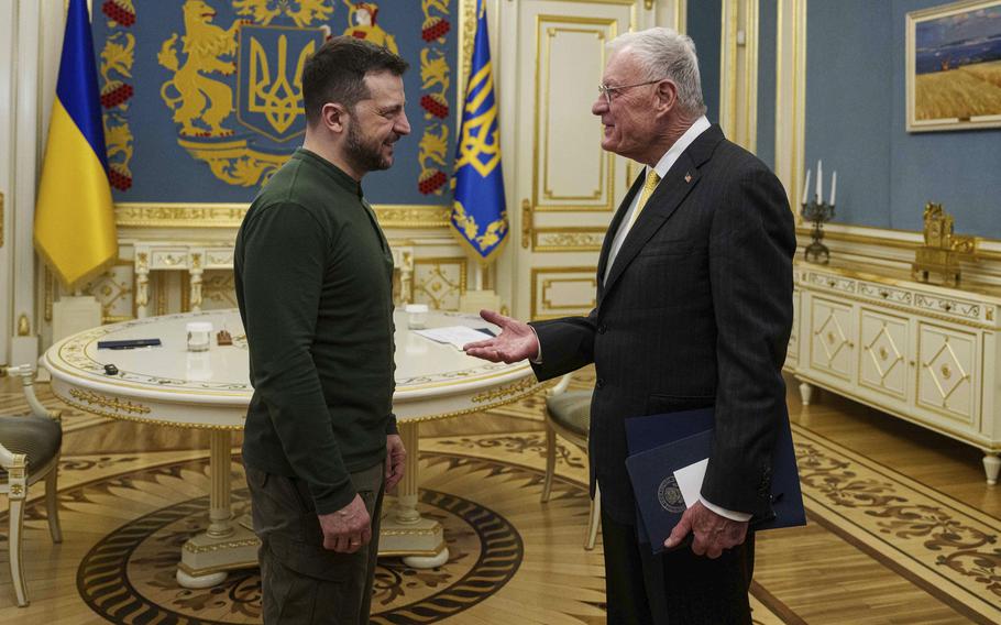 Volodymyr Zelenskyy, wearing a green shirt and pants, and Keith Kellogg, wearing a dark suit, face each other and speak in a Ukrainian government office.