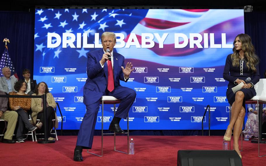 President-elect Donald Trump speaks into a microphone he is holding while seated on a stage, in front of a poster that says “DRILL BABY DRILL.”