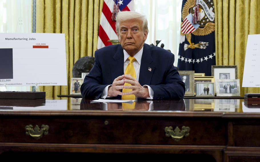 Donald Trump sits at a desk in the Oval Office of the White House with charts about jobs on either side of him. 