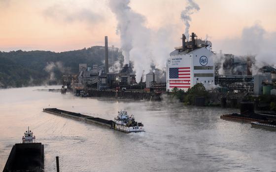 Two tugboats are seen in the water right next to a US Steel plant that has smoke and/or steam billowing out.