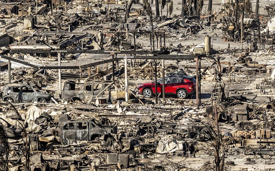 A red car drives among the brown and gray of burned-out homes, trees and vehicles.