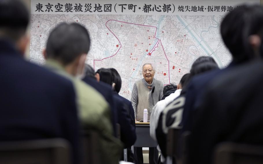 An elderly woman stands in front of a map of Tokyo while speaking to an audience.