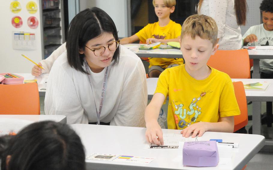 An American elementary school student works on an assignment while a Japanese teacher looks on.