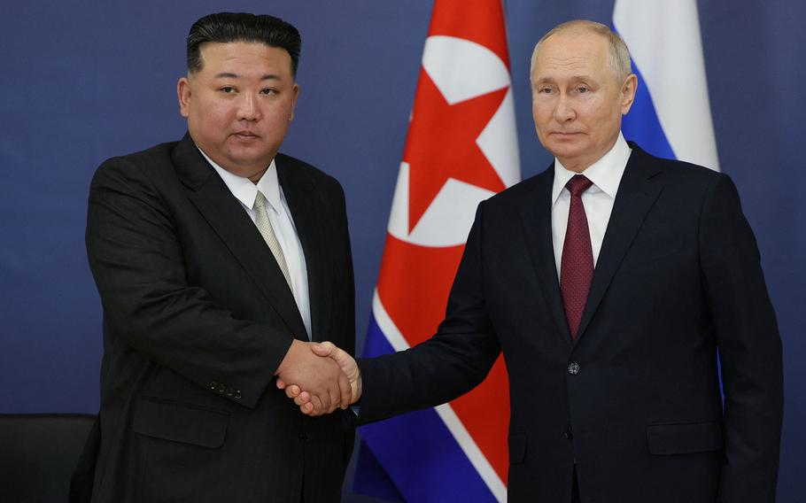 Kim Jong Un and Vladimir Putin shake hands while standing in front of North Korean and Russian flags.