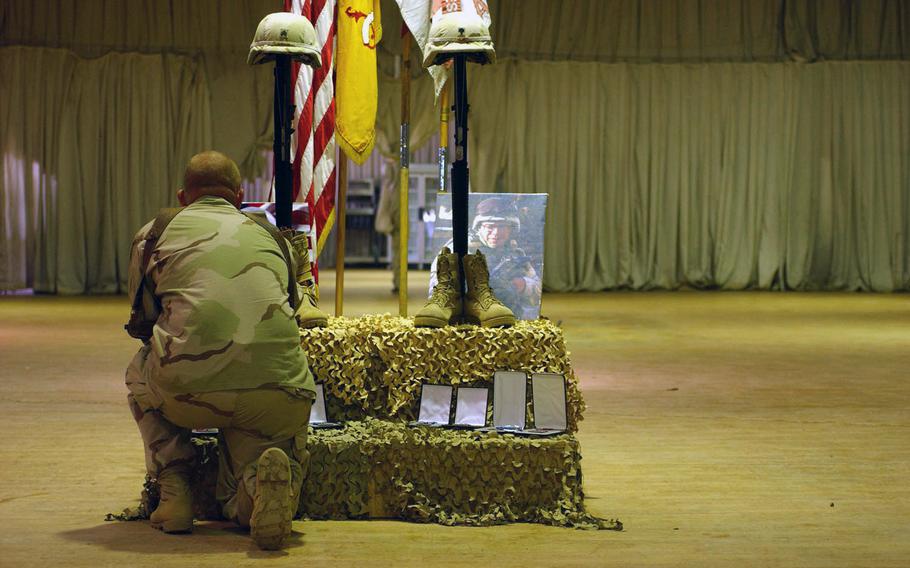 A soldier at Camp Taji kneels in front of a memorial to Sgt. Jonathan Shields and Spc. Jose Valez with boots, helmets and a photograph, flanked by flags.