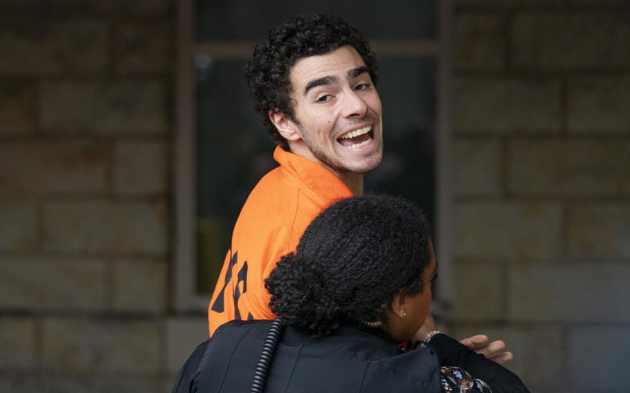 A man in an orange prison jumpsuit looks over his shoulder and speaks while being escorted by a police officer.