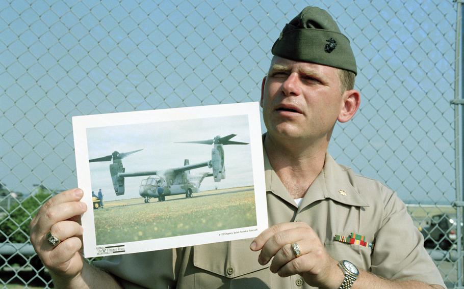 Maj. Barry Moore stands in uniform in front of a chainlink fence, holding a photo of a V-22 Osprey.