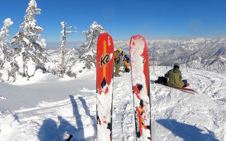 A pair of skis stick out of the snow in the foreground while three people view a mountain vista in the background.