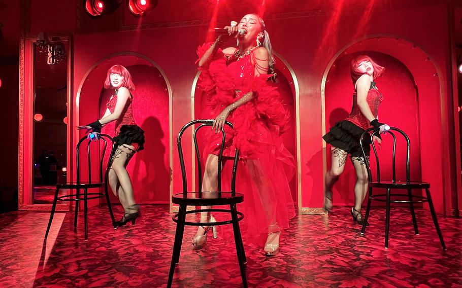 Performers sing and dance around chairs on a stage swathed in red light.