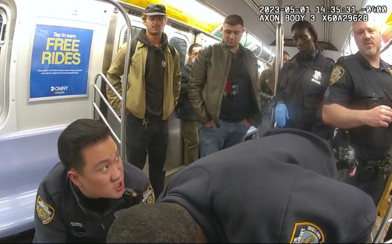 Video still image of police officers attempting to revive a man in a subway car as riders stand and observe.