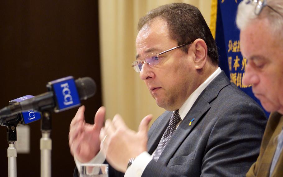 The Ukrainian Ambassador to Japan, wearing a suit and glasses, gestures with his hands while sitting at a table and speaking into a microphone.