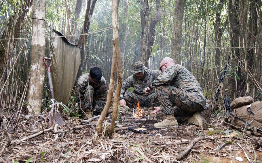 Three Marines hover in brush around a small fire.