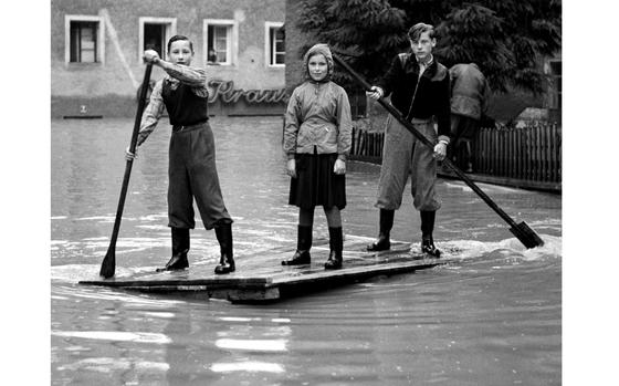 West Germany, July 12, 1954: Although they might resemble Huck, Tom and Becky on a raft excursion on the Mississippi, the sign in the background indicates that these three Bavarian children are caught in the middle of something much less idyllic. Flood waters, rising to the second story of buildings in this town, left more than 50,000 people homeless along the Danube, Isar and Inn rivers. 

Looking for Stars and Stripes’ historic coverage? Subscribe to Stars and Stripes’ historic newspaper archive! We have digitized our 1948-1999 European and Pacific editions, as well as several of our WWII editions and made them available online through https://starsandstripes.newspaperarchive.com/

META DATA: Climate; flooding; weather; 