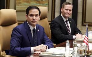 Marco Rubio, wearing a dark blue suit, sits at a conference table with binders of documents and glasses of water in front of him.