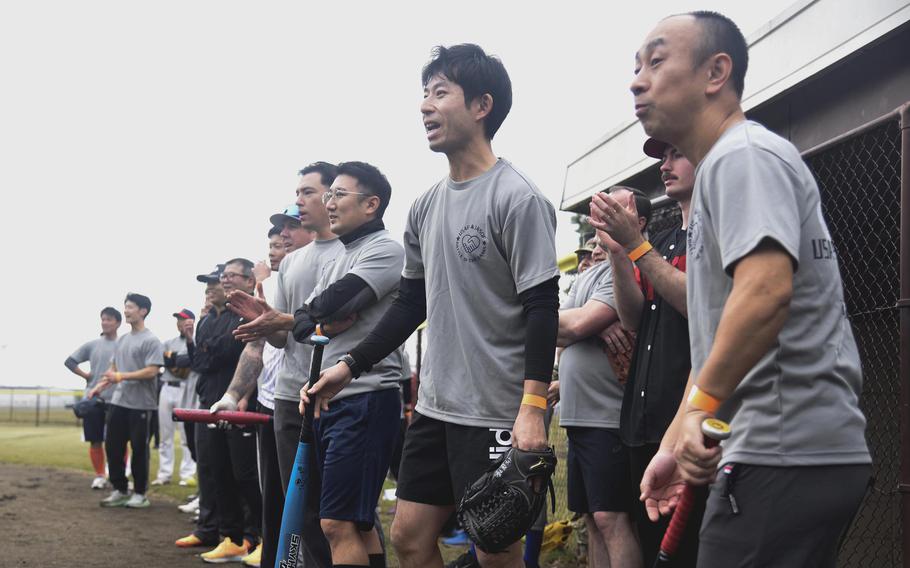 Japan Air Self-Defense Force members wearing grey t-shirts, one holding a blue bat, another one clapping, wear expressions of surprise.