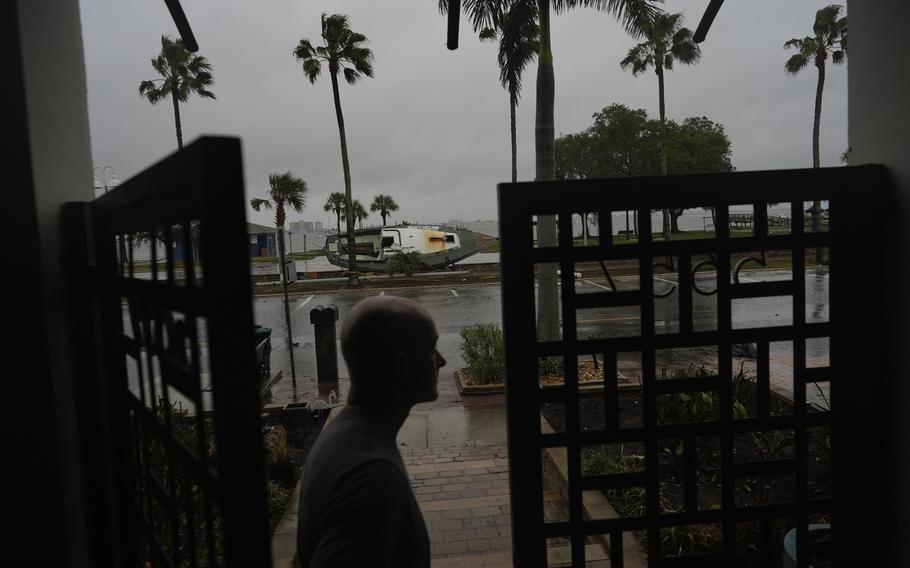 A man stands by the gate of his home with an overturned boat in the background.