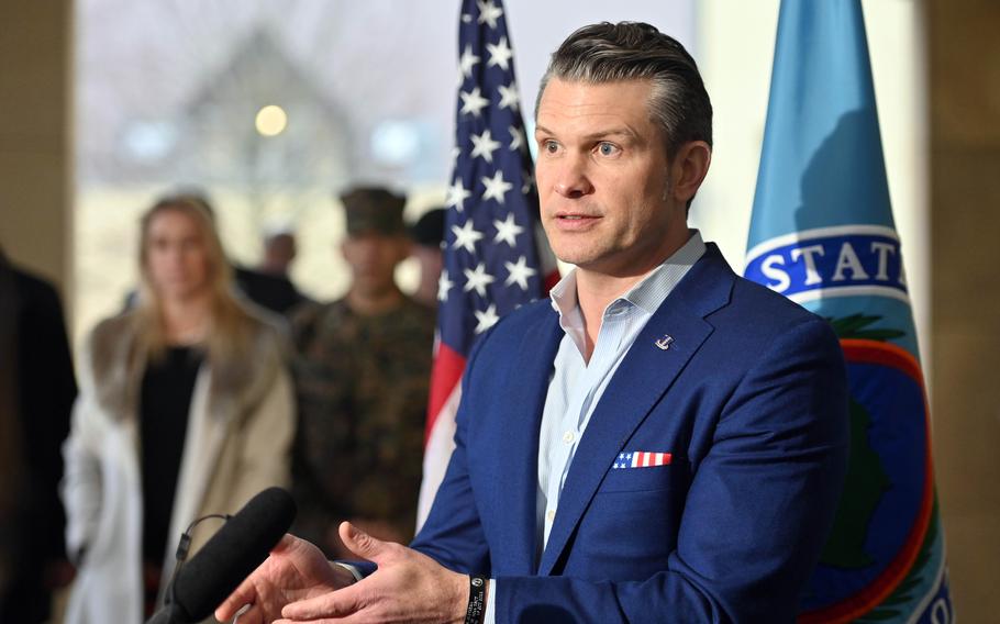 Pete Hegseth holds his hands out while talking, with a U.S. flag in the background