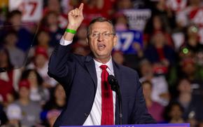 A man in a suit and glasses gestures to the crowd while standing at a podium and speaking into a microphone at a campaign event.