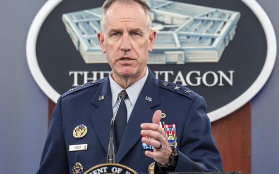 The Pentagon’s press secretary raises one hand in a gesture while seen at a podium from the chest up.