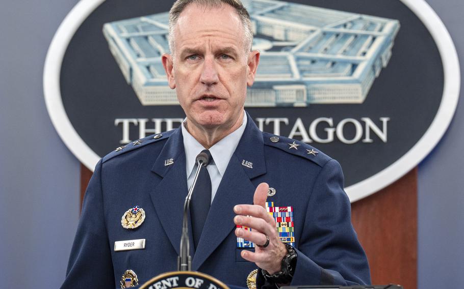 The Pentagon’s press secretary raises one hand in a gesture while seen at a podium from the chest up.