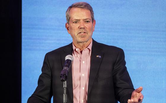 A man in a black suit jacket and red striped shirt speaks in front of a microphone.