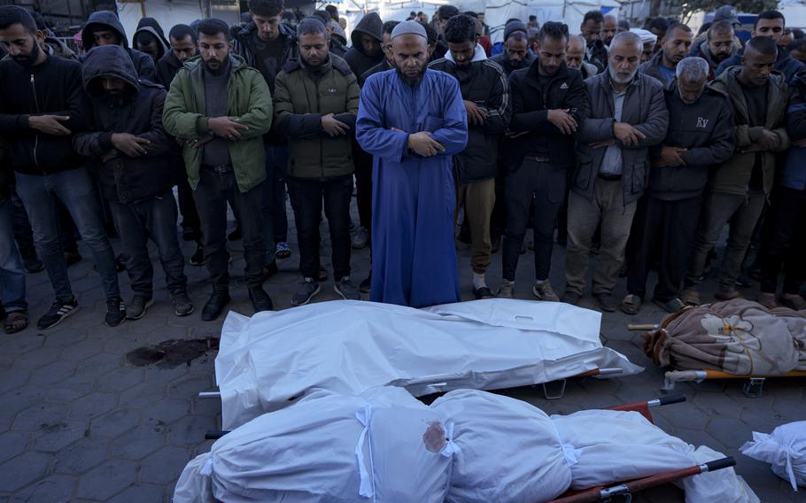 Men stand with their heads bowed in prayer in front of lines of sheet covered corpses.