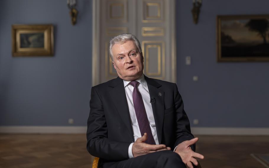 A man in a suit with gray hair sits with his legs crossed, gesturing with his hands in his lap.