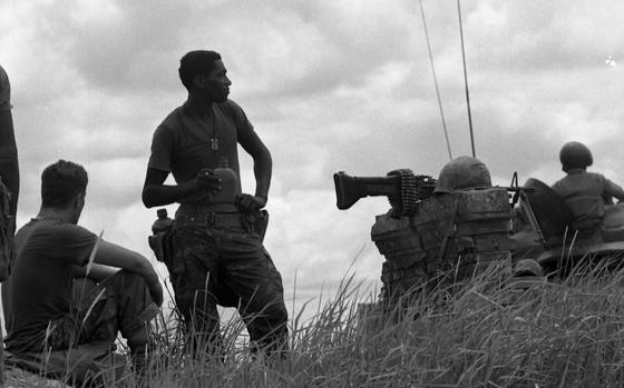 A Marine holds a canteen while standing next to a seated Marine in a grassy field.