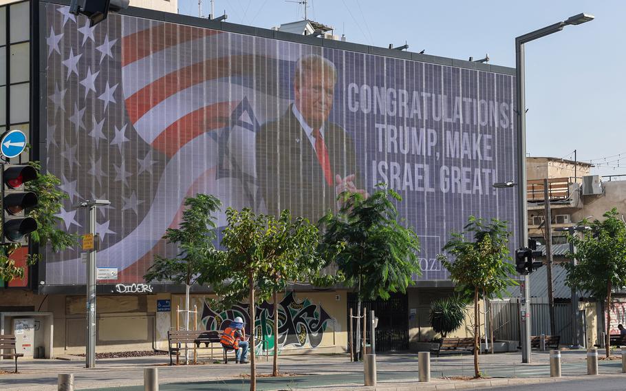 A man sits on a public bench in Tel Aviv beneath a large billboard congratulating Donald Trump.