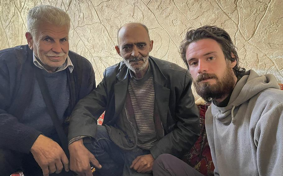 An American man with a beard sits next to two Syrian men in front of a blank wall.