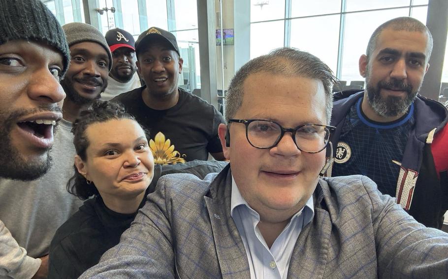 A man wearing black-rimmed glasses and a gray blazer poses with six released American prisoners in front of large windows in an airport.