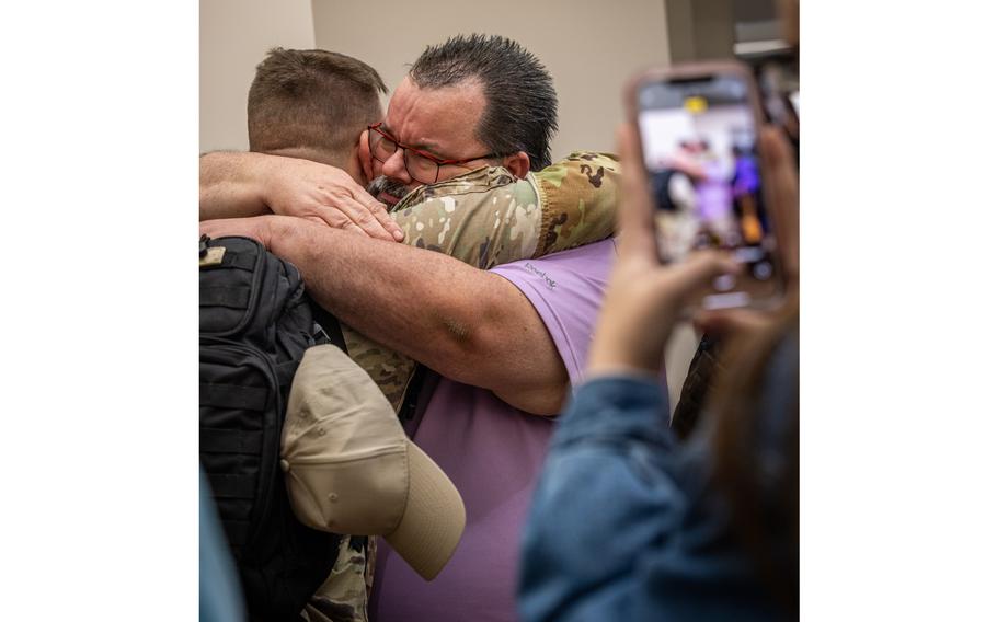 An older man embraces a soldier, seen from behind, while an out-of-focus phone in the foreground takes a picture of the scene.