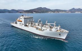 A military ship traverses open water with a mountainous coastline in the background.