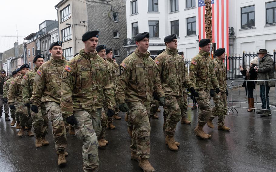 A group of U.S. soldiers marching, with an American flag draped on a building in the background.