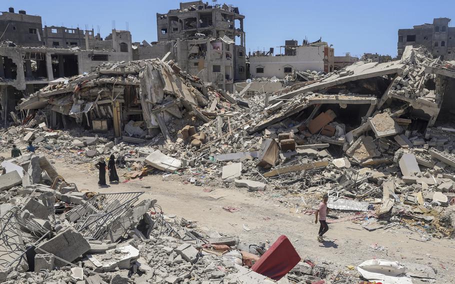 Palestinians walk along a dirt road past huge piles of rubble from destroyed buildings.